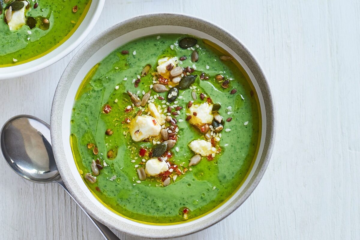 A bowl of chilled green soup topped with crumbled feta cheese and seeds.
