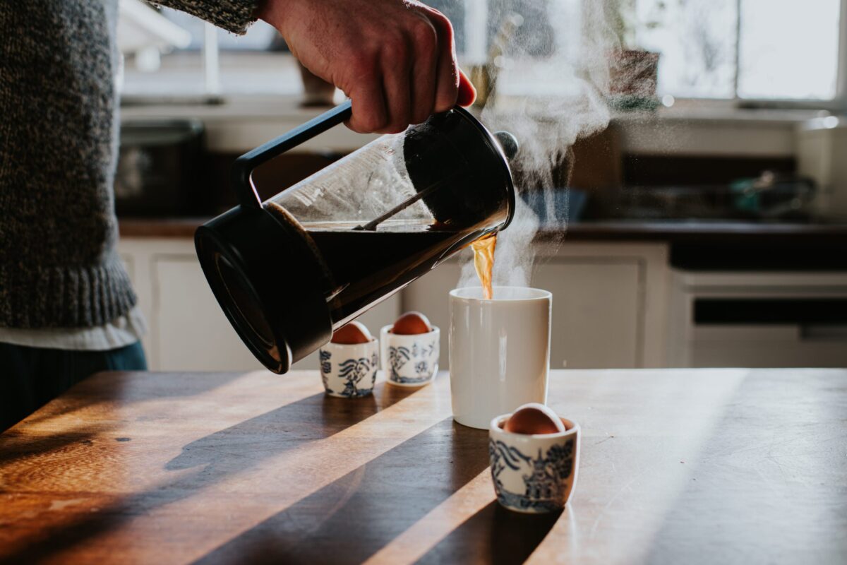 A person pouring coffee from a French press into a mug, with boiled eggs for breakfast nearby.