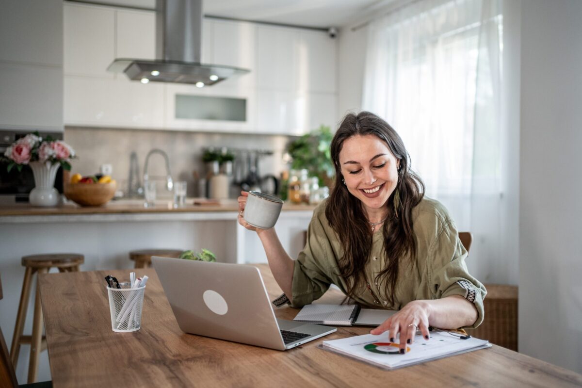 A woman smiling while working from home, enjoying a cup of coffee at her kitchen table.